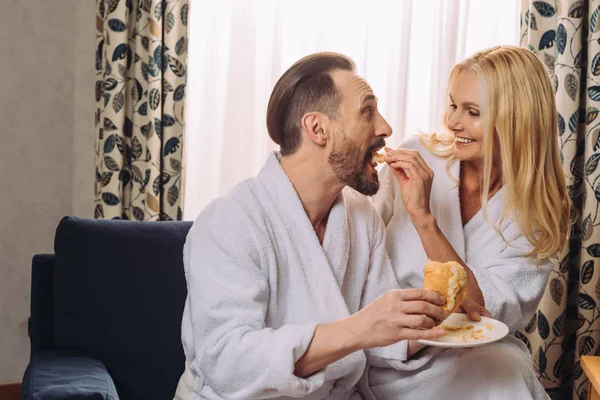 Feliz Mujer Madura Albornoz Alimentar Marido Con Pastelería Durante Desayuno — Foto de stock gratis