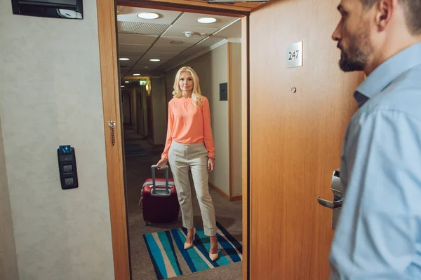 beautiful mature woman with suitcase standing in hallway while bearded man opening door in hotel room