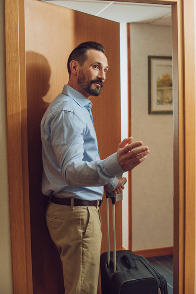 bearded man with suitcase looking away while entering hotel room 