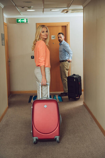 mid adult couple with suitcases looking at camera while walking in hotel hallway