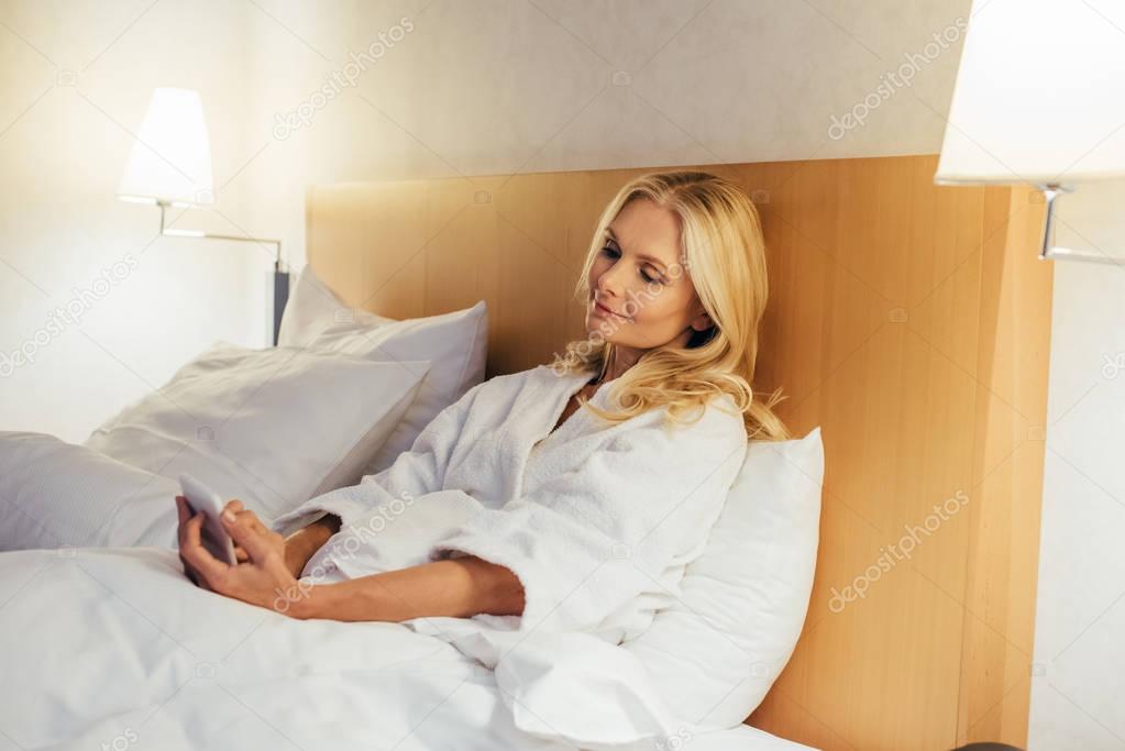 attractive mature woman in bathrobe using smartphone while lying in bed at hotel room 