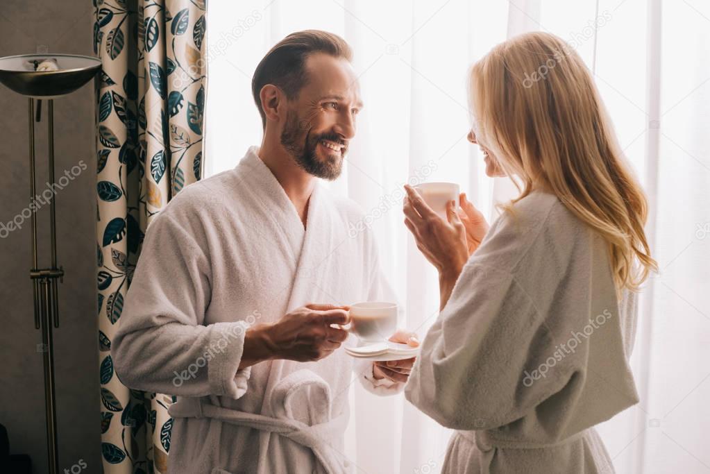 happy middle aged couple in bathrobes drinking coffee and smiling each other in hotel room   