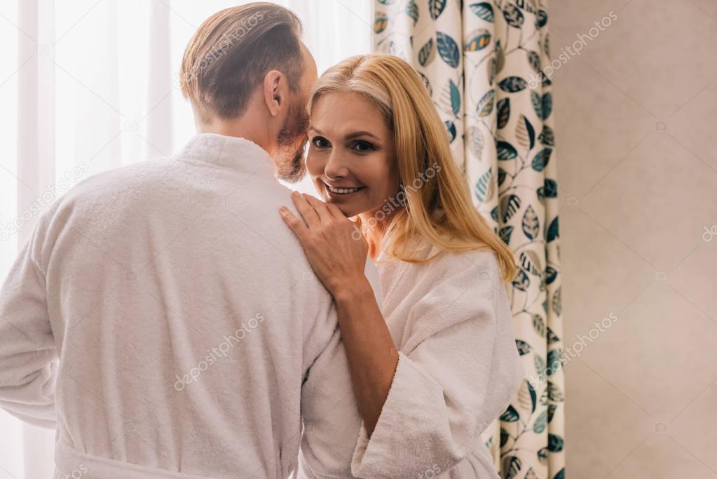 happy mature couple in bathrobes hugging and woman smiling at camera in hotel room 
