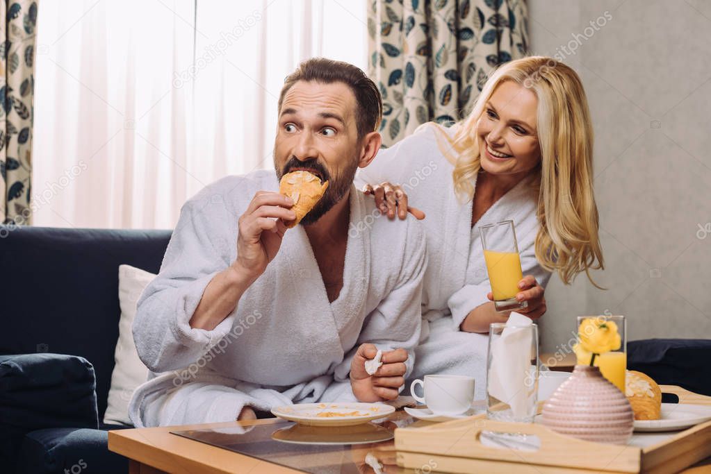 happy mature couple drinking juice and eating pastry during breakfast in hotel room 