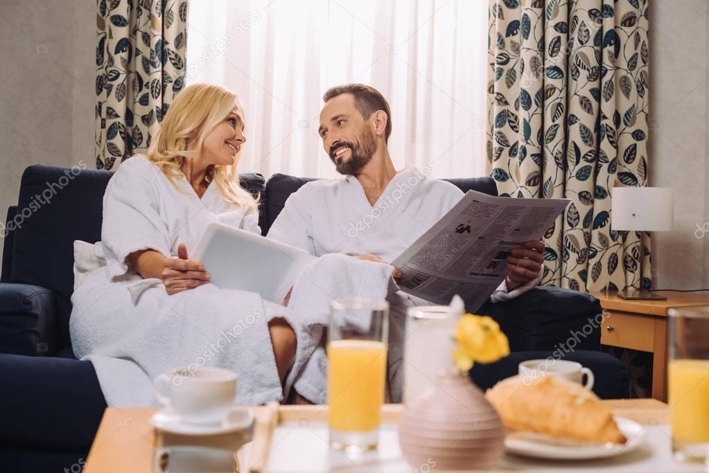 smiling mature couple in bathrobes holding newspaper and digital tablet while having breakfast in hotel room 