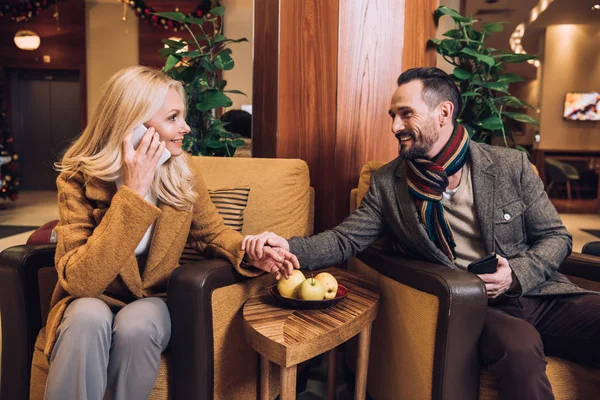 cheerful mature couple holding hands and looking at each other while woman talking on smartphone in hotel