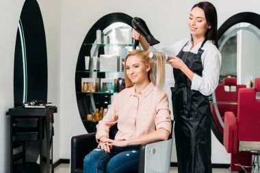 smiling hairdresser drying customer hair at salon clipart