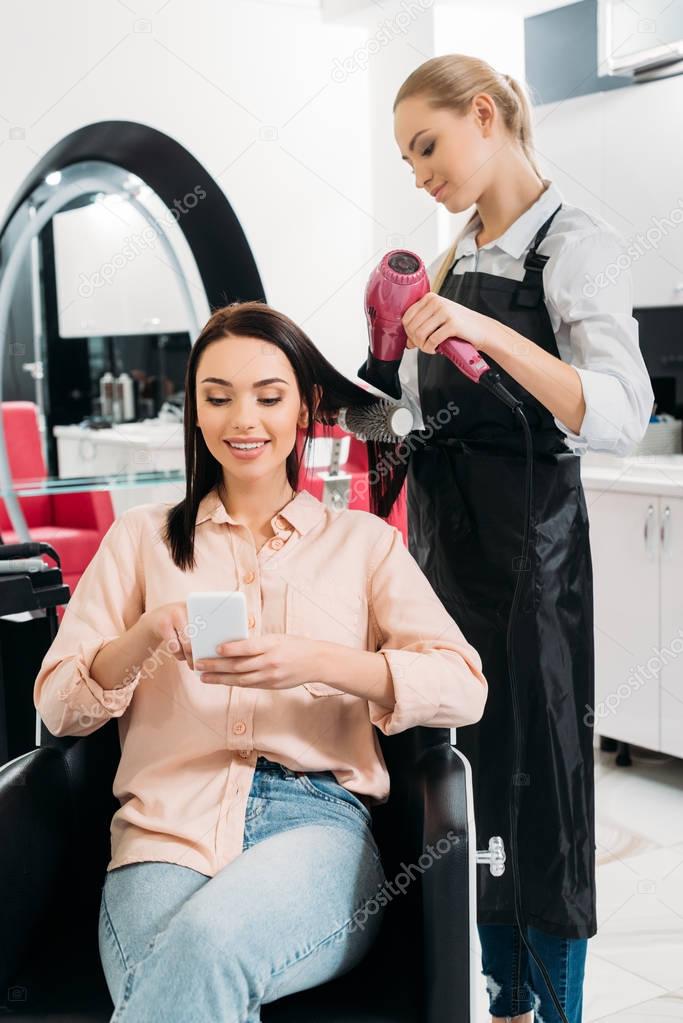  woman looking at smartphone while hairdresser styling hair
