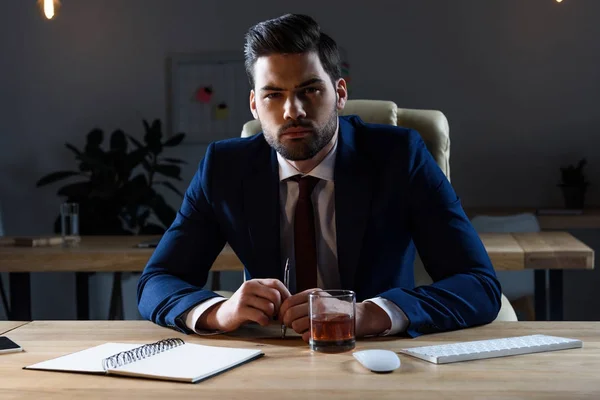 angry businessman sitting at table with glass of whiskey and looking at camera