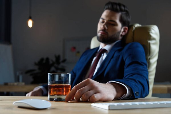 tired businessman sitting in chair and looking at glass of whiskey