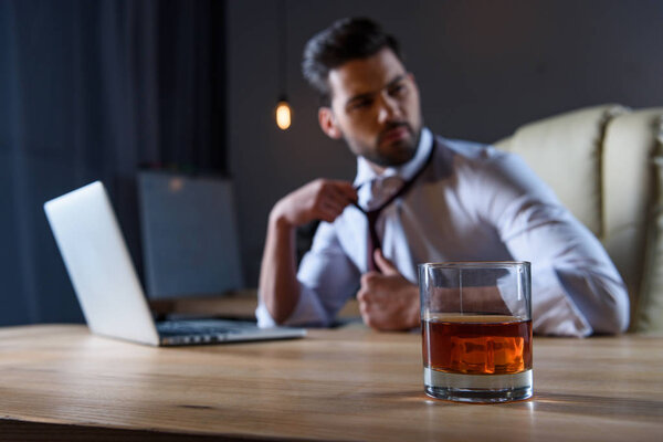 tired businessman loosen tie and sitting at table with glass of whiskey on foreground
