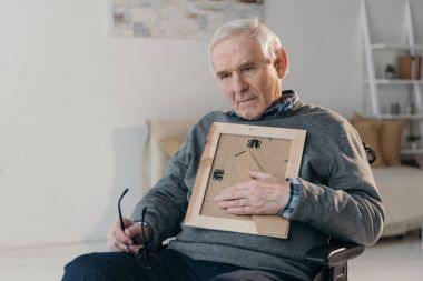 Senior thoughtful man sitting in chair and holding old photo frame clipart