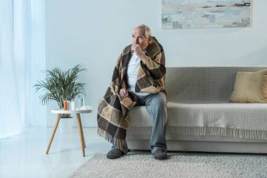 Sick senior man covered in plaid coughs while sitting sofa in room with medications on table clipart