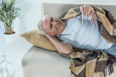Senior sick man covered in plaid lies on sofa and checks thermometer clipart