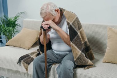 Tired senior man wearing plaid leans on a cane while sitting on sofa clipart