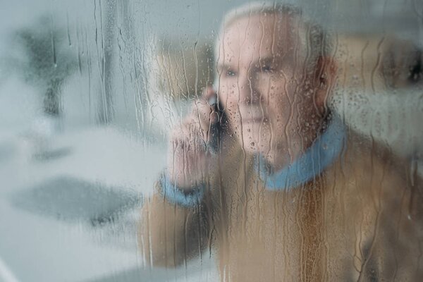 Behind the rainy glass view of senior disturbed man making a phone call 
