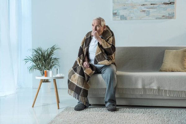 Sick senior man covered in plaid coughs while sitting sofa in room with medications on table