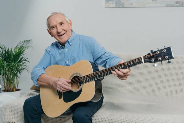Senior happy man plays acoustic guitar while sitting on sofa in room