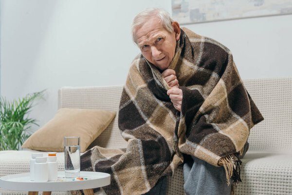 Senior chilled man covered in plaid with medications on table