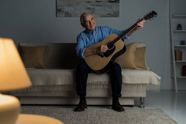 Senior happy man plays acoustic guitar while sitting on sofa in room