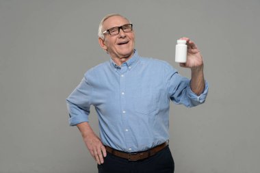Senior smiling man holds white bottle of pills isolated on gray background clipart
