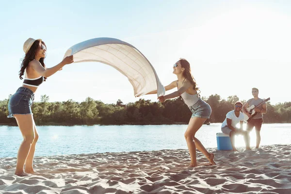 Girls waving beach blanket on riverside — Stock Photo