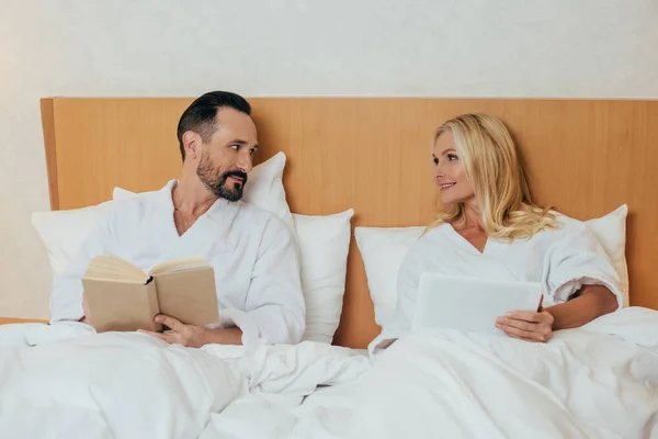 Casal maduro com tablet digital e livro sorrindo uns aos outros enquanto deitado na cama no quarto do hotel — Fotografia de Stock