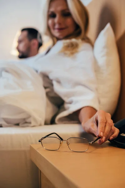 Nahaufnahme einer Frau, die eine Brille auf den Tisch legt, während sie mit ihrem Mann im Hotelzimmer im Bett liegt — Stockfoto