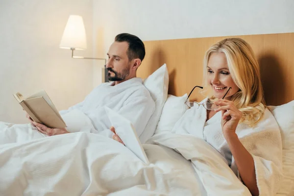 Mulher sorrindo segurando óculos e usando tablet digital enquanto marido lendo livro na cama no quarto do hotel — Fotografia de Stock