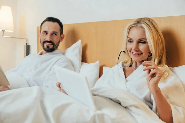 Улыбающаяся зрелая женщина, держащая очки и используя цифровые таблетки, лежа в постели с мужем в гостиничном номере — стоковое фото
