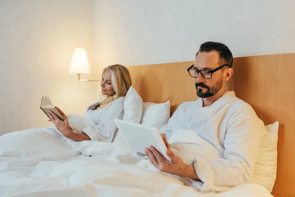 Средняя взрослая пара в халатах читает книги и с помощью цифрового планшета во время лежания в постели в номере отеля — стоковое фото