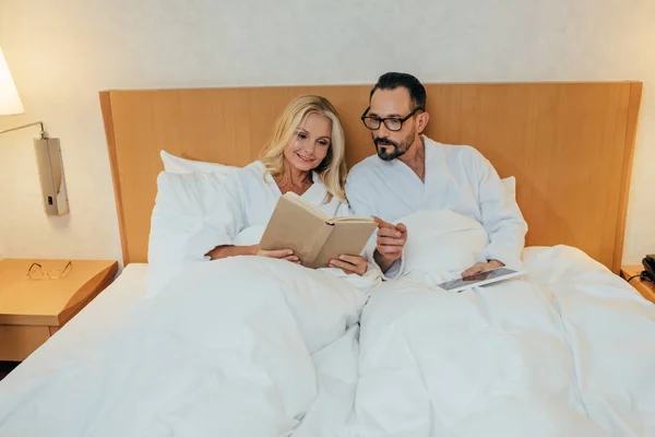 Mature couple in bathrobes reading book and using digital tablet in bed in hotel room — Stock Photo