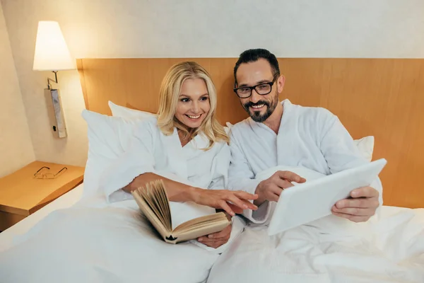 Sonriendo pareja madura leer libro y el uso de tableta digital mientras yacen juntos en la cama en la habitación de hotel - foto de stock