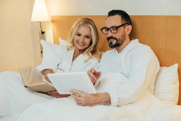 Feliz pareja de mediana edad leer libro y el uso de tableta digital mientras yacen juntos en la cama en la habitación de hotel - foto de stock