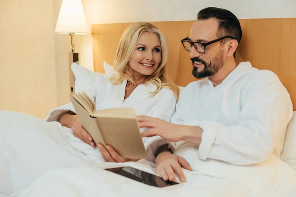 Smiling middle aged couple reading book and using digital tablet while lying together in bed in hotel room — Stock Photo