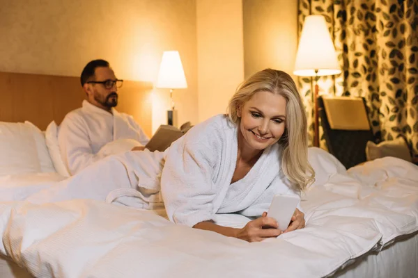 Sorrindo mulher de meia-idade em roupão deitado na cama e usando smartphone enquanto marido lendo livro atrás — Fotografia de Stock