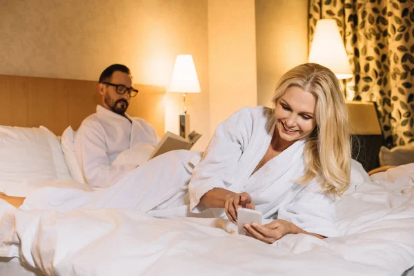 Beautiful smiling middle aged woman in bathrobe lying on bed and using smartphone while husband reading book behind — Stock Photo