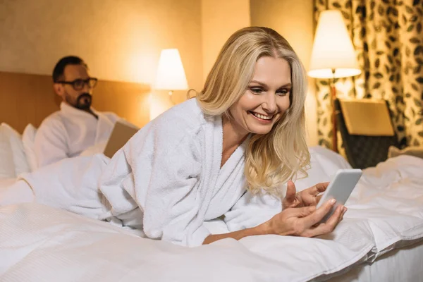 Heureux femme mature en peignoir couché sur le lit et en utilisant smartphone tandis que le mari lecture livre derrière — Photo de stock