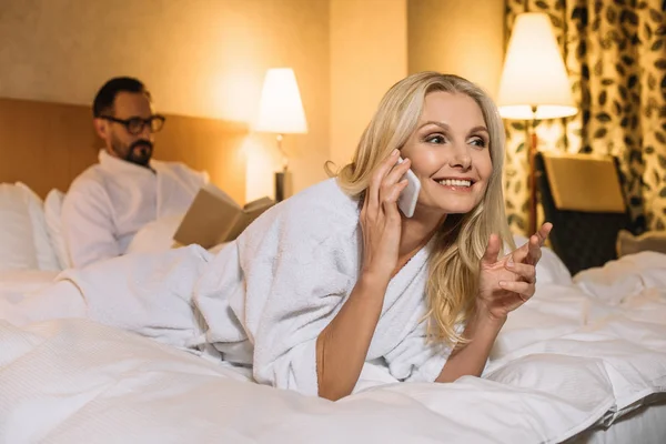 Beautiful mature woman in bathrobe lying on bed and talking on smartphone while husband reading book behind — Stock Photo