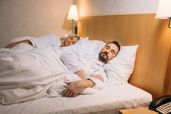 Beautiful middle aged couple in bathrobes sleeping together in bed in hotel room — Stock Photo