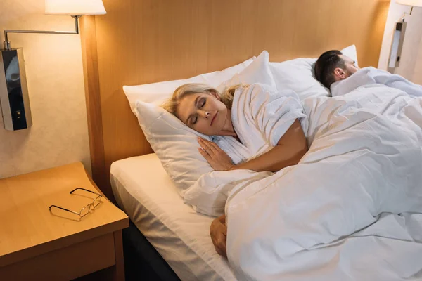 Pareja adulta en albornoces durmiendo en la cama y gafas en la mesa en la habitación de hotel - foto de stock