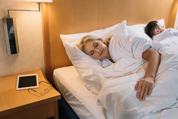 Couple mature en peignoirs dormant au lit et tablette numérique avec des lunettes sur la table dans la chambre d'hôtel — Photo de stock