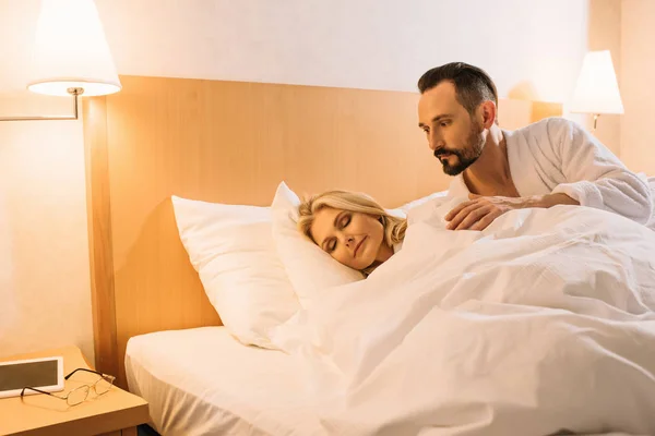 Guapo hombre maduro en albornoz mirando a su esposa durmiendo en la habitación de hotel - foto de stock