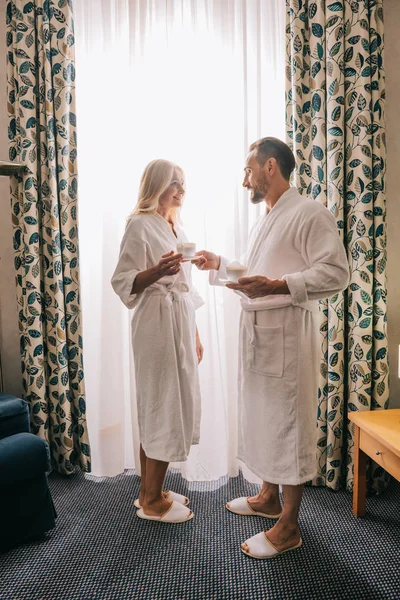 Vista completa de la feliz pareja madura en albornoces bebiendo café y sonriéndose en la habitación del hotel - foto de stock