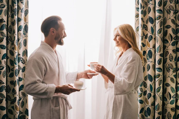 Vista lateral de la feliz pareja de mediana edad en albornoces bebiendo café y sonriéndose en la habitación del hotel - foto de stock