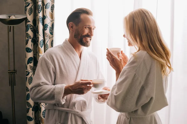 Feliz pareja de mediana edad en albornoces bebiendo café y sonriéndose en la habitación del hotel - foto de stock