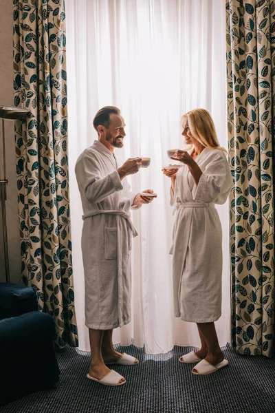 Vista lateral de feliz pareja madura en albornoces bebiendo café y sonriéndose en la habitación del hotel - foto de stock