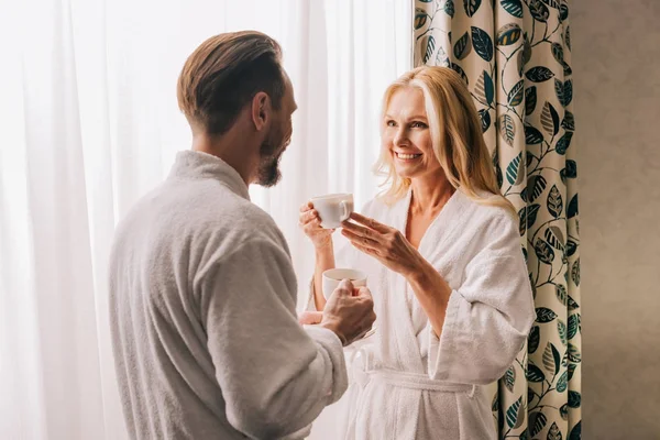 Beautiful happy mature couple in bathrobes drinking coffee and smiling each other in hotel room — Stock Photo