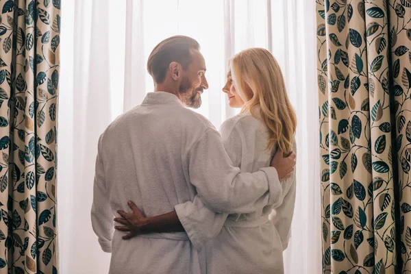 Visão traseira do casal de meia idade no amor vestindo roupões de banho e sorrindo uns aos outros no quarto de hotel — Fotografia de Stock