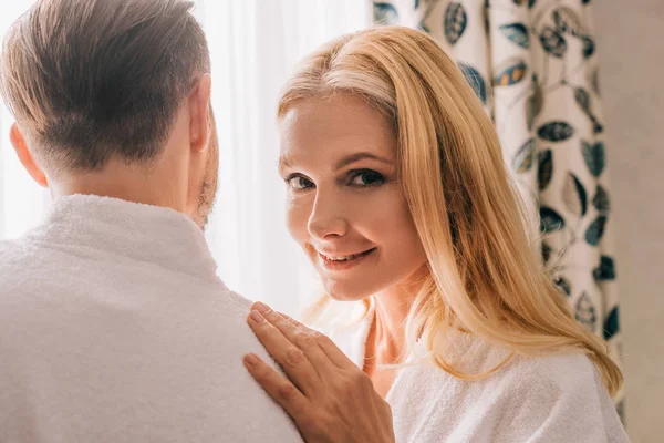 Belle femme mature souriant à la caméra tout en embrassant mari en peignoir dans la chambre d'hôtel — Photo de stock